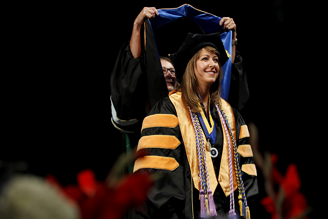 A student on stage at graduation in doctoral robes being presented with a hood over their head