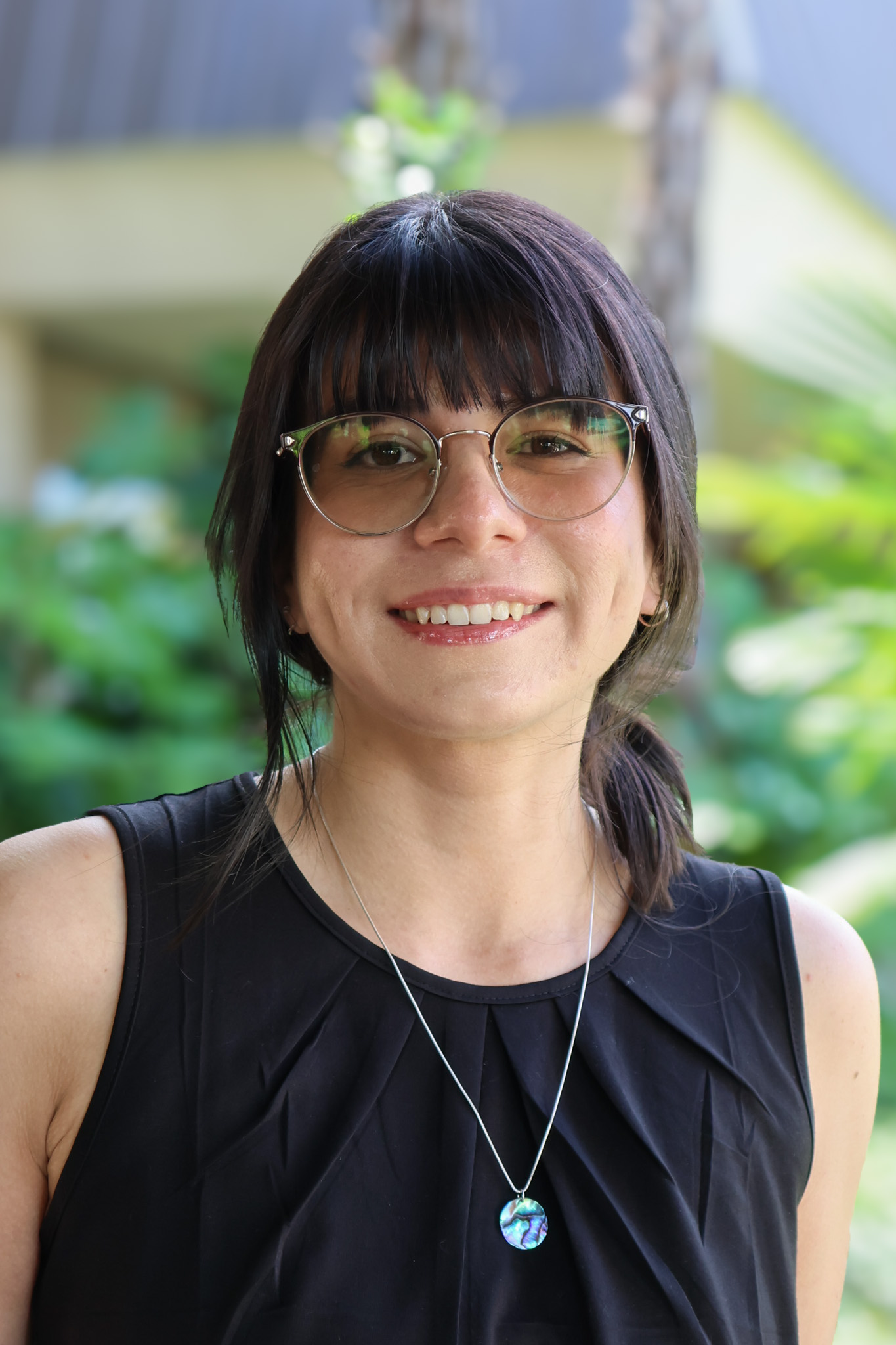 A woman with dark hair and glasses, smiling, wearing a black dress and a silver necklace.