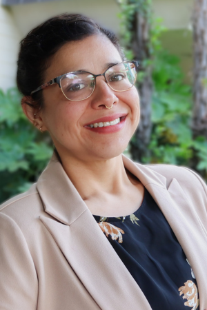 A woman wearing glasses, a black floral dress, and a tan blazer, standing outdoors with greenery in the background.