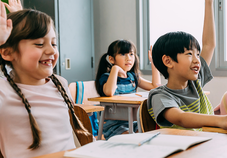 Three school-age students are sitting in class, with their hands raised.